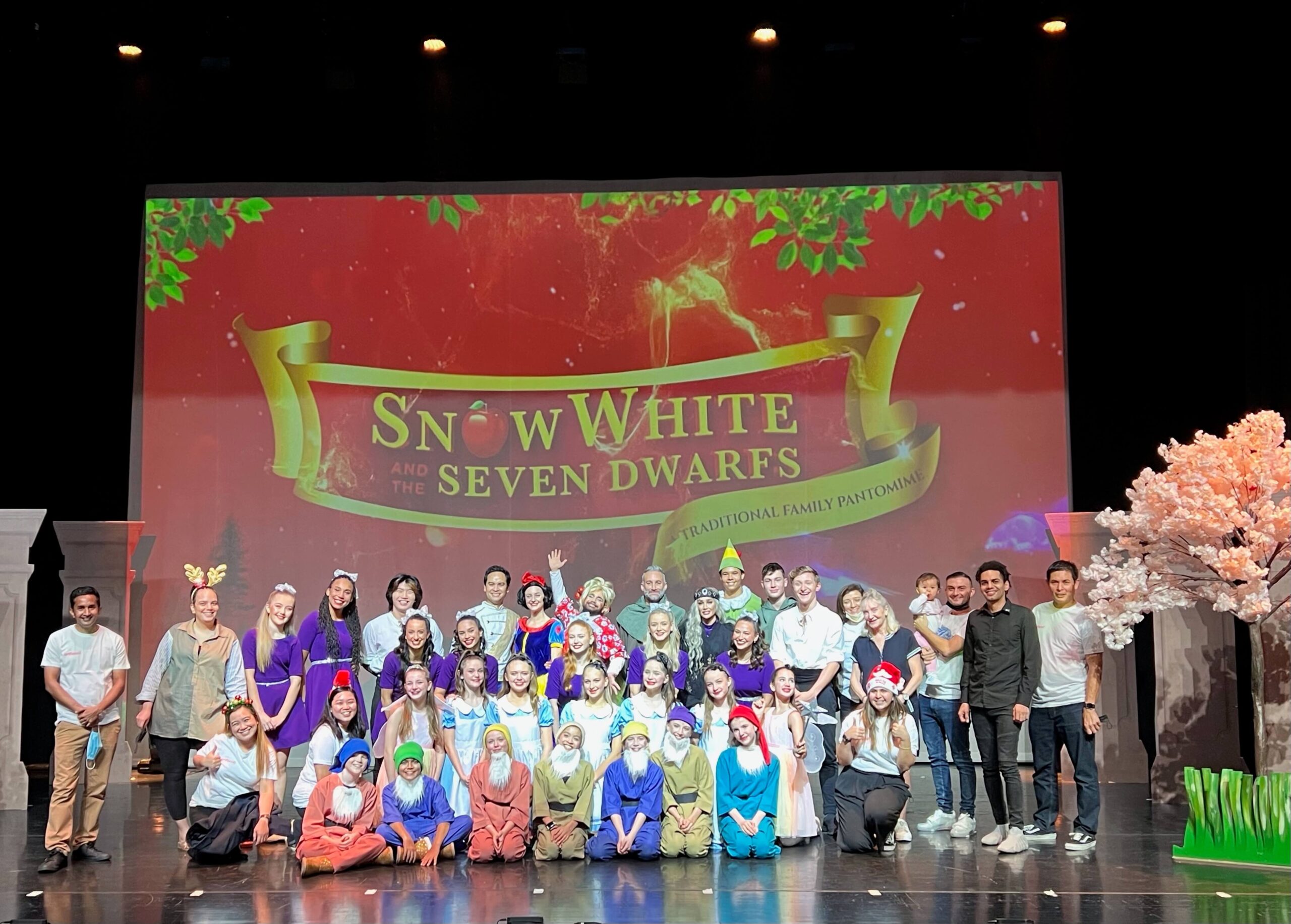 A happy cast photo of a production of Snow White smile on stage with a large projection screen and set pieces while in costumes