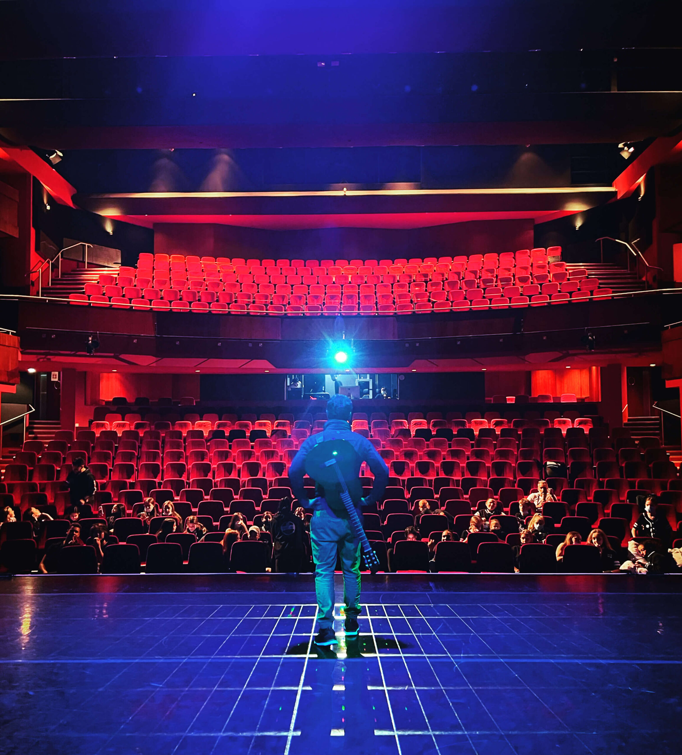 A man stands on a stage with a guitar on his back in front of young students in the audience of one of the Theatres in Dubai