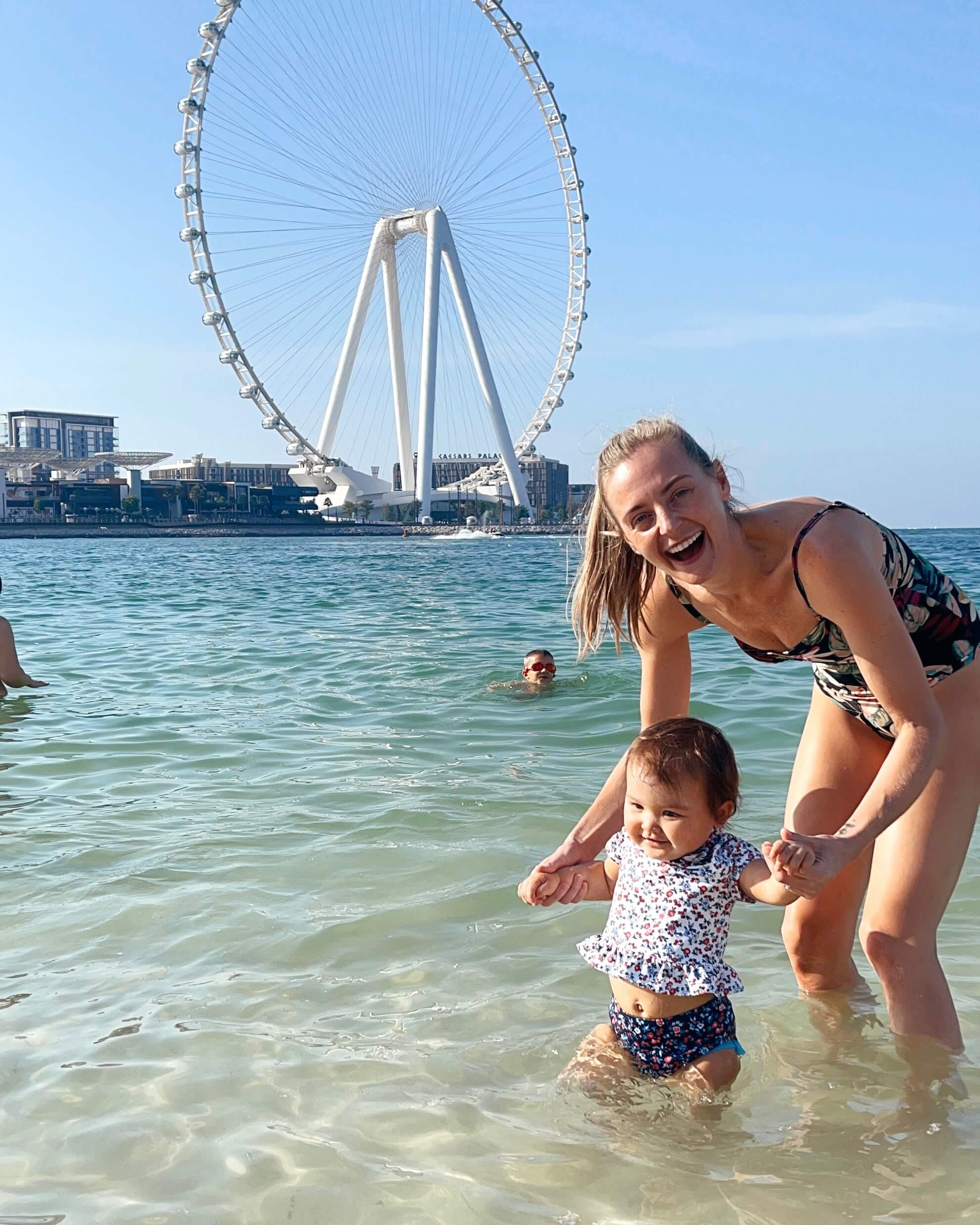 A laughing mom helps her baby walk in shallow beach water by a ferris wheel during family activities in Dubai