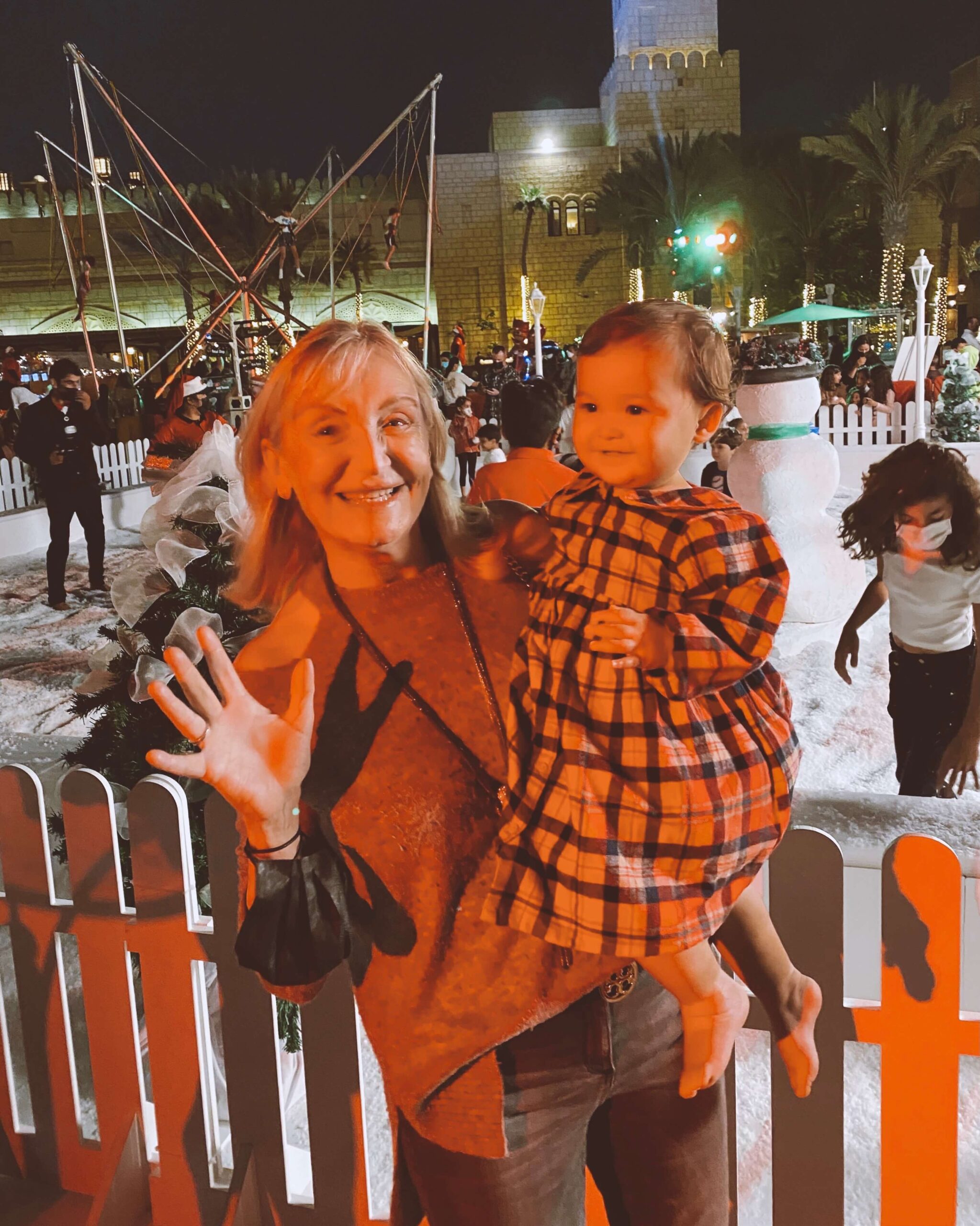 A joyful grandma holds her granddaughter on her hip while exploring a snow filled park at night during a christmas festival