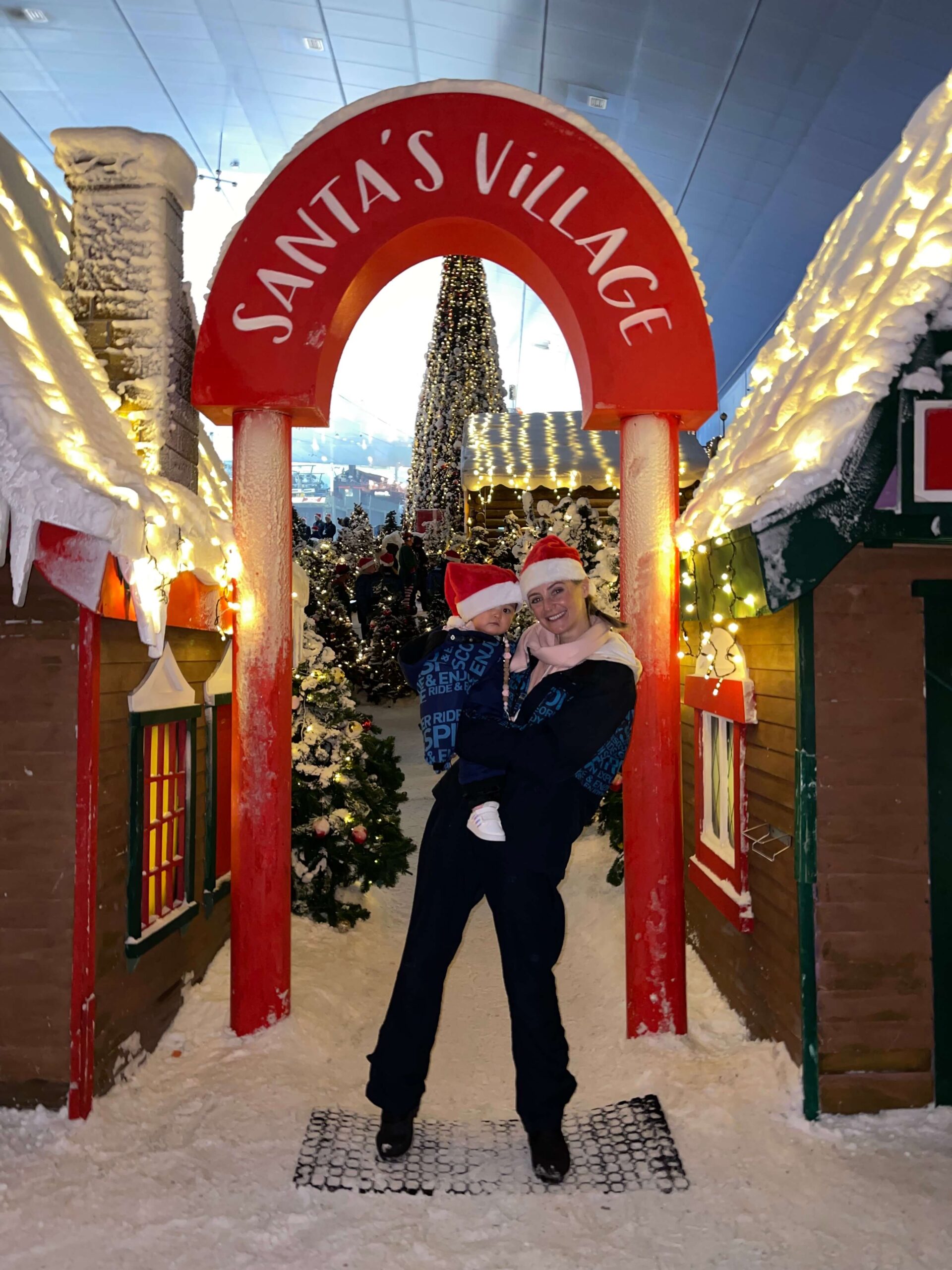 A happy woman stands under a Santa's Village sign with her baby on her hip both in christmas hats