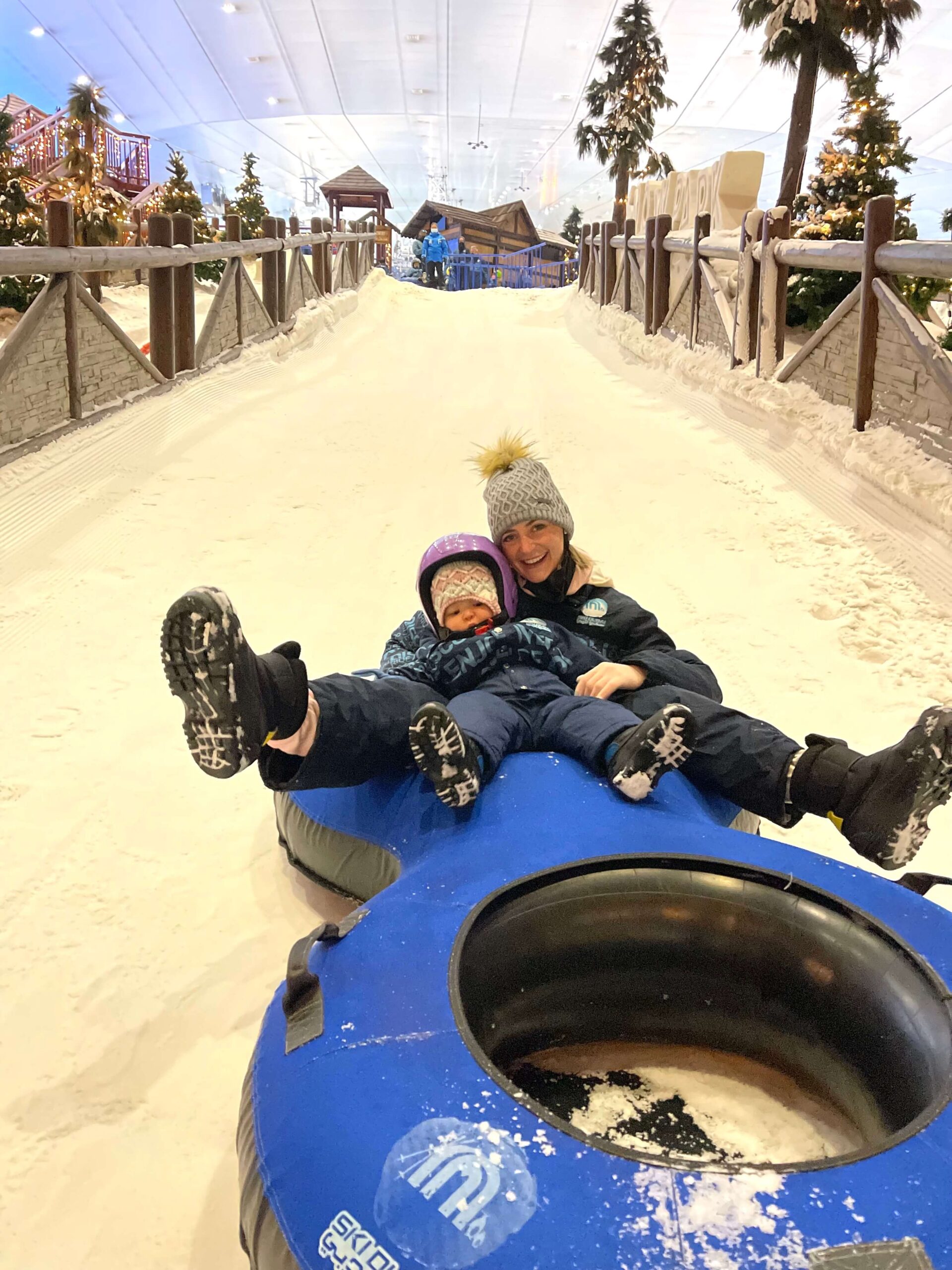 A blonde woman sits in the back of a double person tube on a hill with her baby in her lap at Ski Dubai