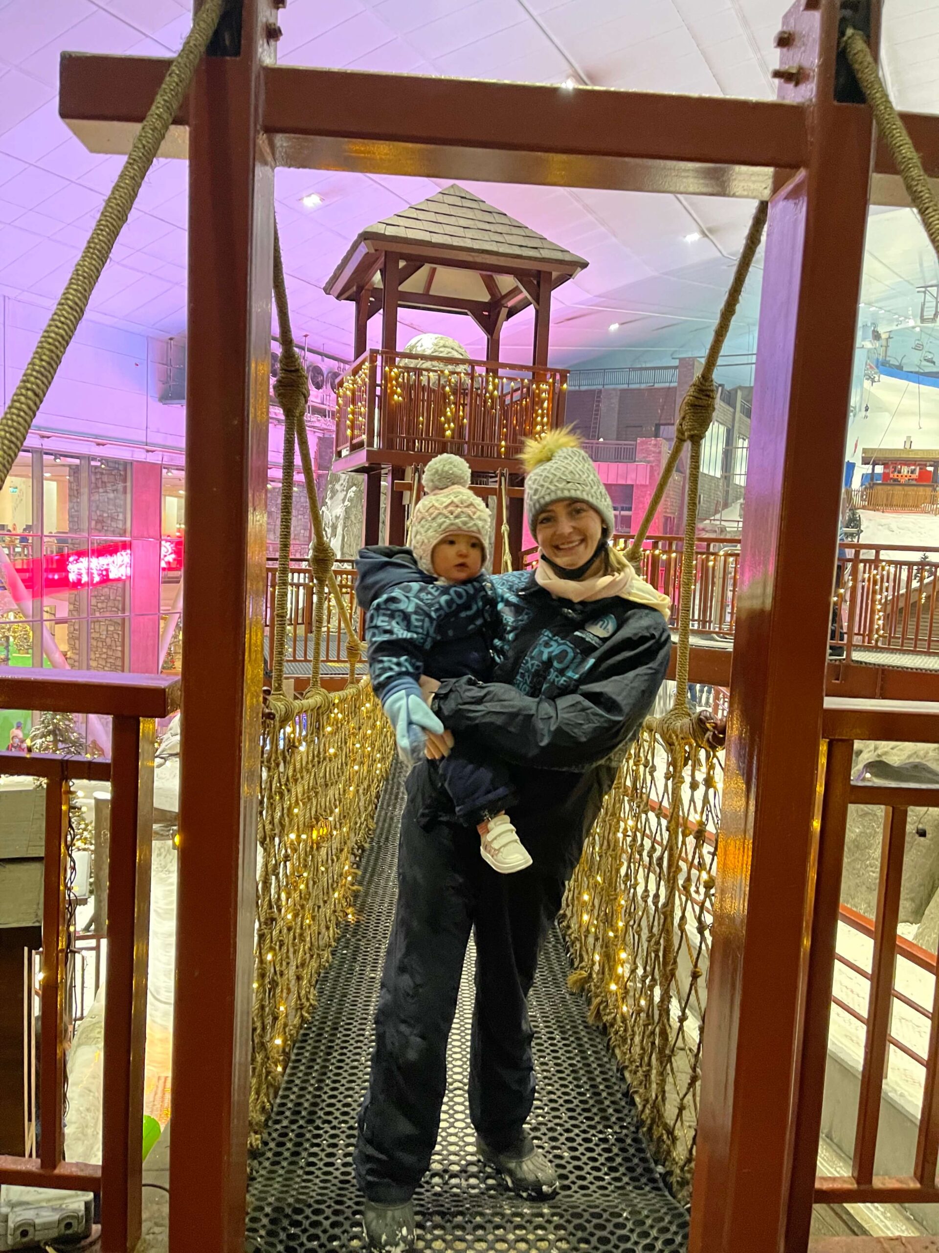 A happy woman in a ski suit stands on a rope bridge holding her baby on her hip at Ski Dubai